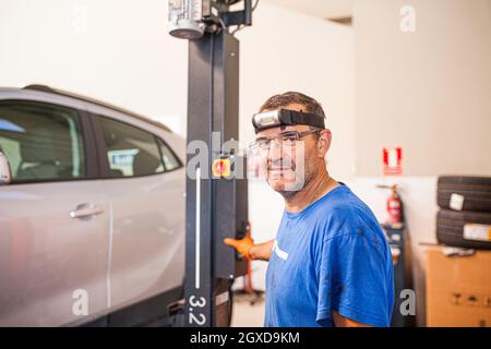 Side view of cheerful mature male technician operating elevator with modern automobile in repair service and looking at camera Stock Photo