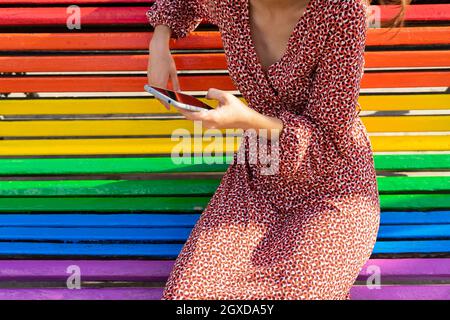 Crop unrecognizable female in dress sitting on colorful bench and browsing mobile phone in daytime Stock Photo