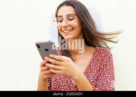 Smiling female in dress and eyeglasses standing near white wall and using smartphone in daytime Stock Photo