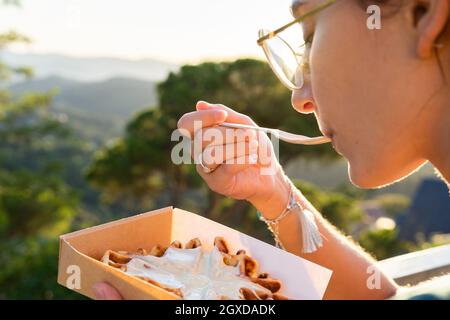 Side view of crop young female eating tasty Belgian waffles with whipped cream in takeaway box against mounts in back lit Stock Photo