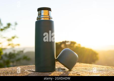 Modern stainless steel and plastic thermos with cup on rough surface under shiny sky at sundown Stock Photo