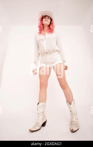 From below of young female with pink hair in stylish white clothes and hat standing in light room Stock Photo