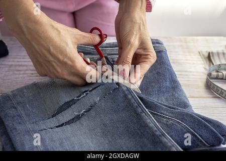 Crop anonymous female seamstress using scissors while cutting jeans on wooden table in atelier Stock Photo