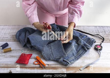 Crop anonymous female seamstress using scissors while cutting jeans pocket on wooden table in atelier Stock Photo