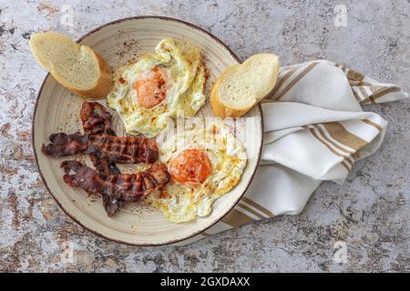 Overhead view of tasty sunny side up eggs with fried bacon strips and wheat bread pieces on plate above towel Stock Photo