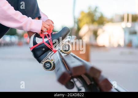 Side view of cropped unrecognizable young female wearing light pink hoodie and black trousers lacing up roller skates leaning leg on back of wooden be Stock Photo