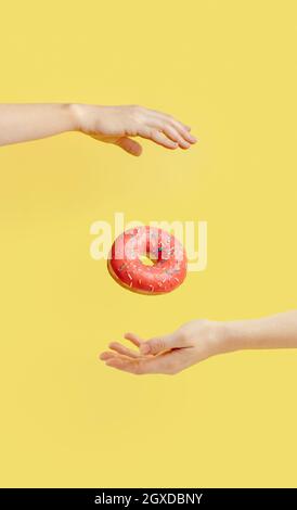 Donut falls between hands on bright yellow paper background. Donuts delivery, diet, sugar concept. Female hands and glazed donut with sprinkles flying Stock Photo