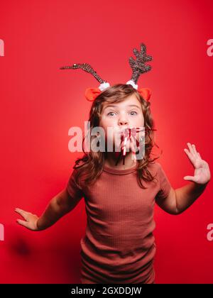 Joyful little girl in casual clothes and festive deer headband blowing party whistle and looking at camera during Christmas celebration against red ba Stock Photo