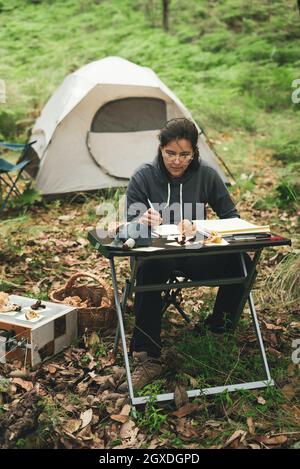 Thoughtful female in casual clothes and eyeglasses sitting at table and taking notes in notebook while researching mushrooms in green park against ten Stock Photo