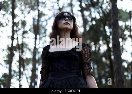 From below mystic witch in long black dress and with painted face standing looking away in dark gloomy woods Stock Photo