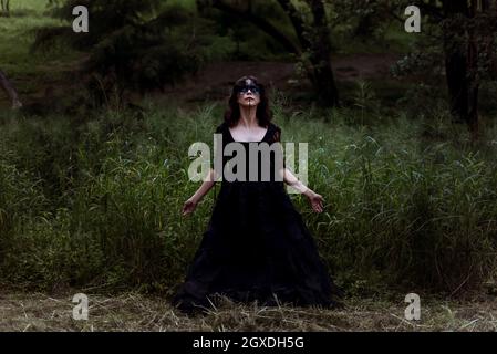 Mystic witch in long black dress and with painted face standing looking up in dark gloomy woods Stock Photo