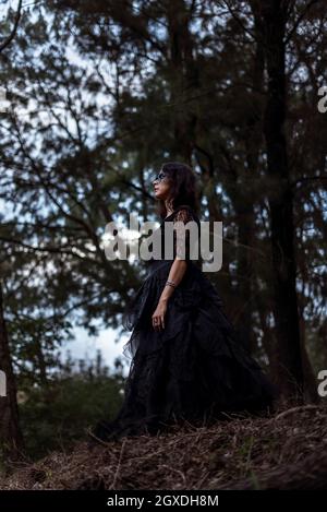 From below mystic witch in long black dress and with painted face standing looking away in dark gloomy woods Stock Photo