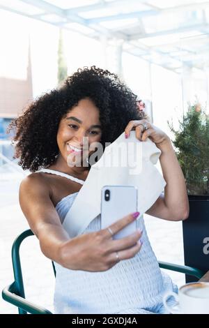 Positive pregnant African American female sitting at table with laptop and latte and taking self portrait with baby clothes Stock Photo