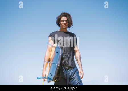 Young male athlete with curly hair and tattoos in wet sportswear holding surfboard while looking at camera under light sky Stock Photo