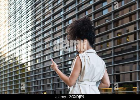 Back view of young African American female with curly hair watching cellphone while carrying netbook past modern building Stock Photo