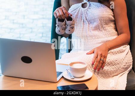 Cropped unrecognizable African American female sitting at table with cup of latte and showing baby booties to netbook during video call Stock Photo