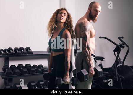 Strong sportsWoman in activewear standing with dumbbells near muscular bodybuilder during workout in gym in daytime Stock Photo