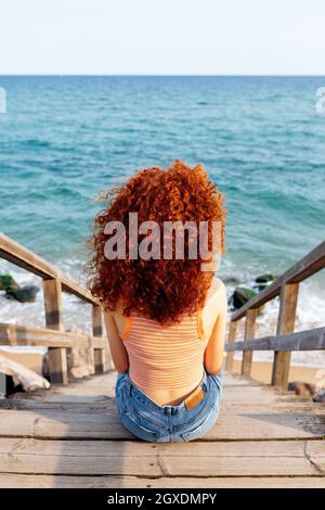 High angle back view of unrecognizable female traveler with long curly ginger hair sitting on wet sandy beach washed by foamy splashing waves Stock Photo