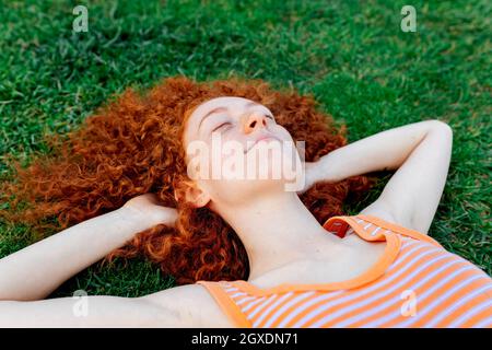 Top view of happy curly haired female with hands behind head relaxing on lawn with eyes closed Stock Photo
