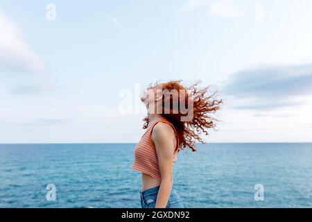 Serious young blond haired slim female tourist touching hat with eyes closed while lounging in sunlight under blue cloudless sky Stock Photo