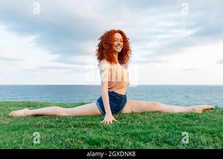 Delighted red haired female with curls doing Hanumanasana for stretching legs on grassy coast of rippling sea and looking at camera Stock Photo
