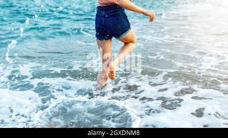 Barefoot cropped unrecognizable female traveler running along sandy beach washed by foamy waves in windy weather Stock Photo