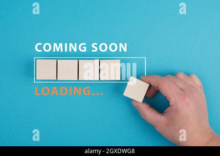 The words coming soon are standing over a loading bar, hand puts the last wooden cube to the line, blue colored background Stock Photo