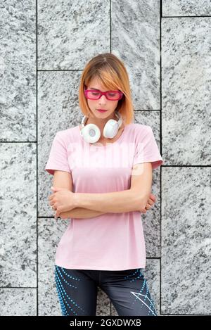 Thoughtful young female in stylish pink sunglasses with headphones on neck dressed in sporty t shirt and pants standing with arms crossed against gray Stock Photo