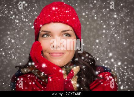 Happy Mixed Race Woman Wearing Winter Hat and Gloves Enjoys Watching the Snow Fall Looking to the Side on Gray Background. Stock Photo