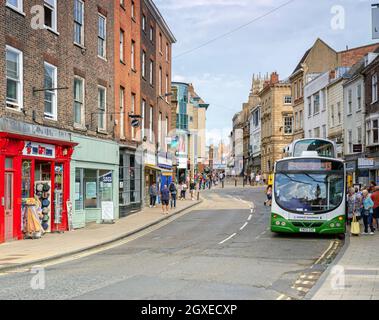 A city street with a bus and a queue of people at the door.  Shops line the pavements and people walk around. A sky with cloud is above. Stock Photo