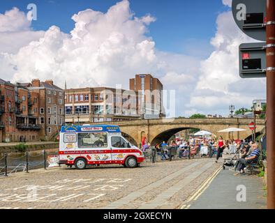 A stone bridge spans a river with buildings on the other side.  People are sitting and an ice-cream vendor is beside the river. Stock Photo