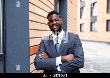 Portrait of confident black businessman in suit posing with crossed arms by office center and smiling at camera Stock Photo