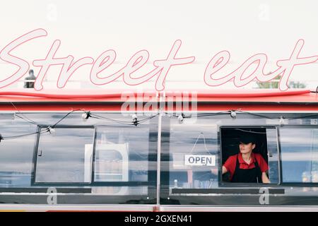 Young woman in uniform hanging opening notice on window of food truck in the morning Stock Photo
