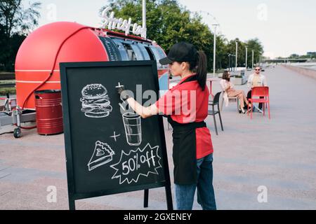Young businesswoman drawing picture of fast food and drink on discount on blackboard Stock Photo