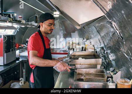 Young clerk of food truck frying snacks in large amount of oil or fat in street truck Stock Photo