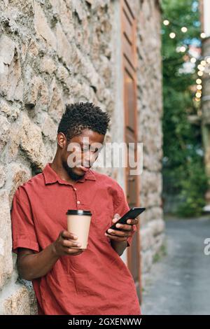 African guy with glass of coffee watching online video in smartphone outdoors Stock Photo