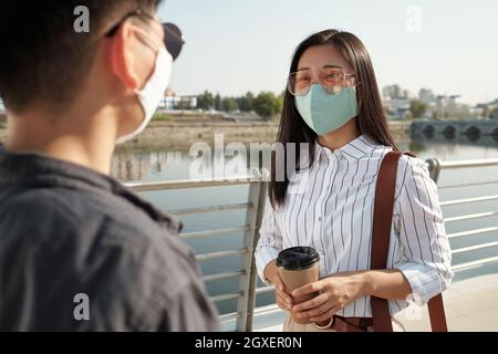 Young Asian female in eyeglasses and protective mask talking to colleague Stock Photo