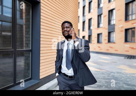 Excited african american businessman talking on smartphone, walking outdoors near modern office center, copy space Stock Photo
