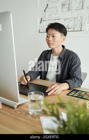 Young Chinese designer with stylus pen drawing on tablet display in front of computer Stock Photo