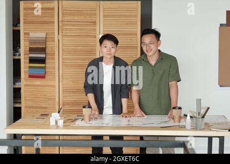 Two young Chinese architects in casualwear looking at you while working over sketch in office Stock Photo