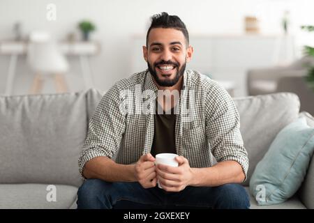 Young smiling Arab guy drinking hot aromatic coffee on sofa at home, copy space Stock Photo