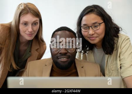 Three happy young employees looking at screen of laptop in front of camera Stock Photo