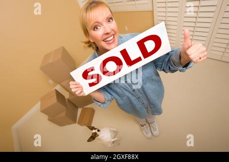 Excited Woman with Thumbs Up and Doggy Holding Sold Real Estate Sign Near Moving Boxes in Empty Room Taken with Extreme Wide Angle Lens. Stock Photo