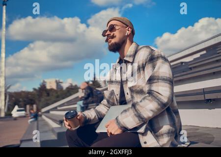 Young man listens music on his headphones sits outdoor while holds laptop and coffee in hands Stock Photo
