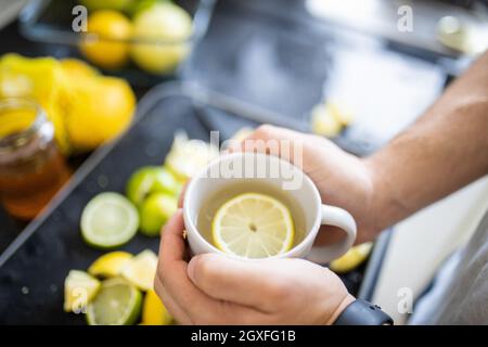 Male hands holding cup of lemon tea above black tray with slices of limes and lemons. Man with cup of tea with lemon slice floating inside. Sweet citr Stock Photo