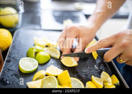 Male hands carefully slicing lemons and limes above black tray. Man cutting lemons into small pieces on a tray. Citrus drink preparation Stock Photo