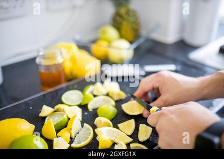 Male hands carefully slicing lemons and limes above black tray. Man cutting lemons into small pieces on a tray. Citrus drink preparation Stock Photo
