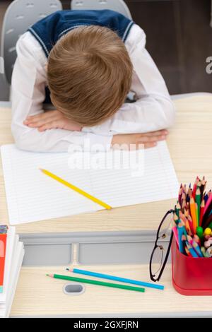 A cute first grader boy in a school uniform at home during a pandemic fell asleep doing homework at a desk with books and pencils. Selective focus. Cl Stock Photo