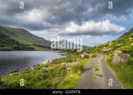 Winding country road leading trough Black Valley, between lake and rocky hills of MacGillycuddys Reeks mountains, Ring of Kerry, Ireland Stock Photo