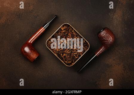 Close up tin full of ready rubbed long coarse cut tobacco blend and two handmade briar smoking pipes over grunge brown background, elevated top view, Stock Photo